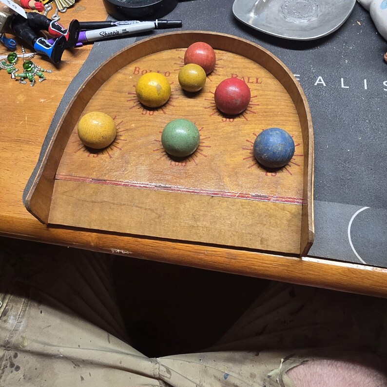 K&ouml;nnte beinhalten: Ein Vintage-Holz-Ballwurfspiel mit einer gebogenen R&uuml;ckwand und mehreren farbigen B&auml;llen. Die B&auml;lle sind gelb, rot, gr&uuml;n und blau. Das Spielbrett hat Punktwerte unterhalb der Position jedes Balls.