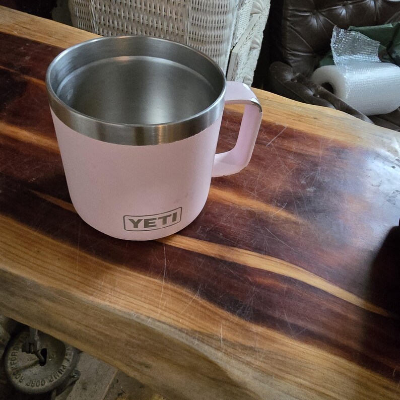 K&ouml;nnte beinhalten: Ein rosa isolierter Becher mit Edelstahl-Innenraum und Griff. Auf der Vorderseite des Bechers ist der Markenname "YETI" aufgedruckt. Der Becher steht auf einer Holzoberfl&auml;che.