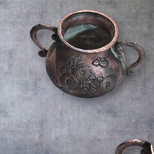 Op de afbeelding: Een antieke koperen theepot met twee handvatten en een bloemenontwerp. De theepot heeft een verweerde, gepatineerde uitstraling en is versierd met reli&euml;fbloemen.