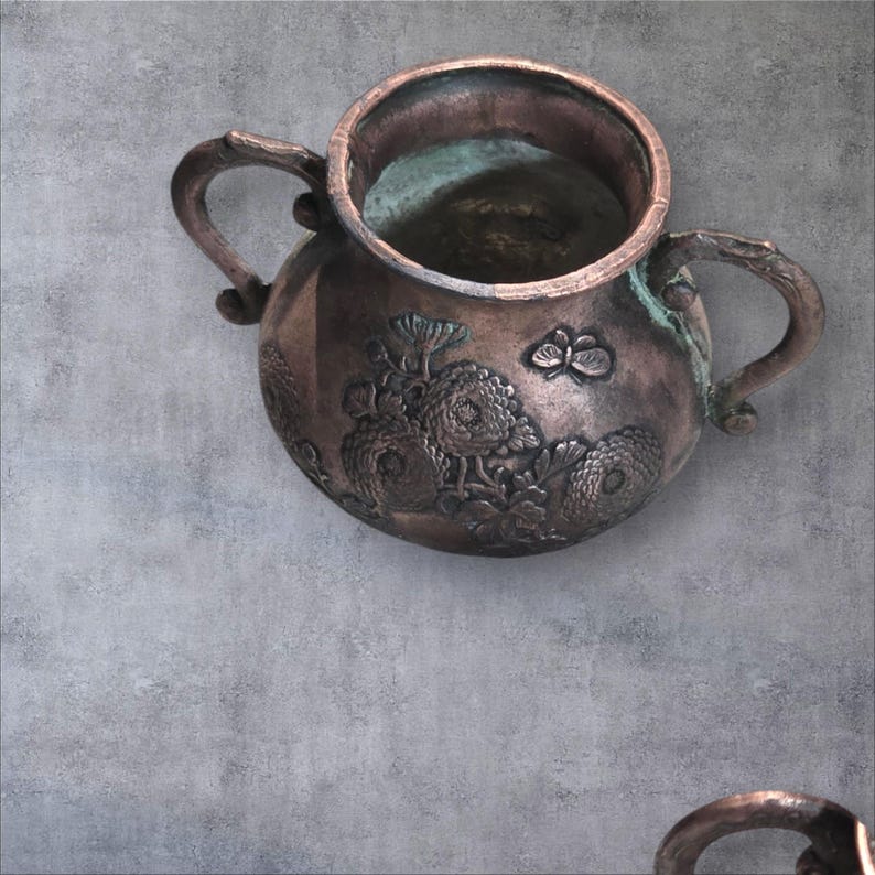 Op de afbeelding: Een antieke koperen theepot met twee handvatten en een bloemenontwerp. De theepot heeft een verweerde, gepatineerde uitstraling en is versierd met reli&euml;fbloemen.