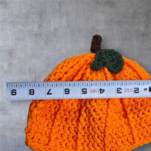 Puede incluir: Sombrero de calabaza de punto de ganchillo naranja con un tallo y una hoja de fieltro verde. El sombrero mide aproximadamente 15 cm de di&aacute;metro.