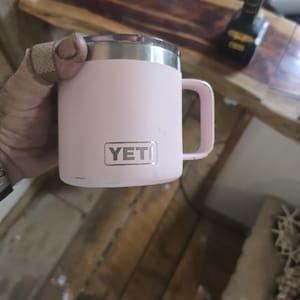 K&ouml;nnte beinhalten: Ein rosa isolierter Becher mit silbernem Rand und Henkel. Der Becher hat den Markennamen "YETI" in einem rechteckigen silbernen Etikett. Der Becher ist f&uuml;r hei&szlig;e oder kalte Getr&auml;nke konzipiert.