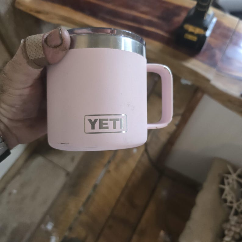 K&ouml;nnte beinhalten: Ein rosa isolierter Becher mit silbernem Rand und Henkel. Der Becher hat den Markennamen "YETI" in einem rechteckigen silbernen Etikett. Der Becher ist f&uuml;r hei&szlig;e oder kalte Getr&auml;nke konzipiert.