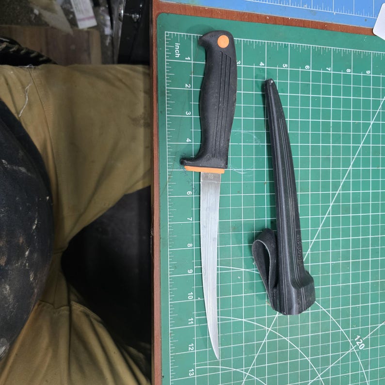 May include: A black and orange handled fillet knife with a silver blade, approximately 13 inches long, next to a black sheath. The knife is on a green cutting mat.