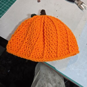 Puede incluir: Un gorro de calabaza tejido a crochet en color naranja con un tallo verde. El gorro est&aacute; hecho con un punto texturizado y tiene una forma redondeada.