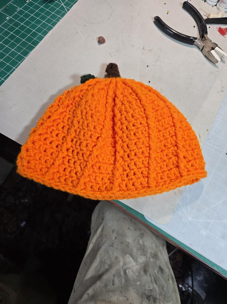Puede incluir: Un gorro de calabaza tejido a crochet en color naranja con un tallo verde. El gorro est&aacute; hecho con un punto texturizado y tiene una forma redondeada.