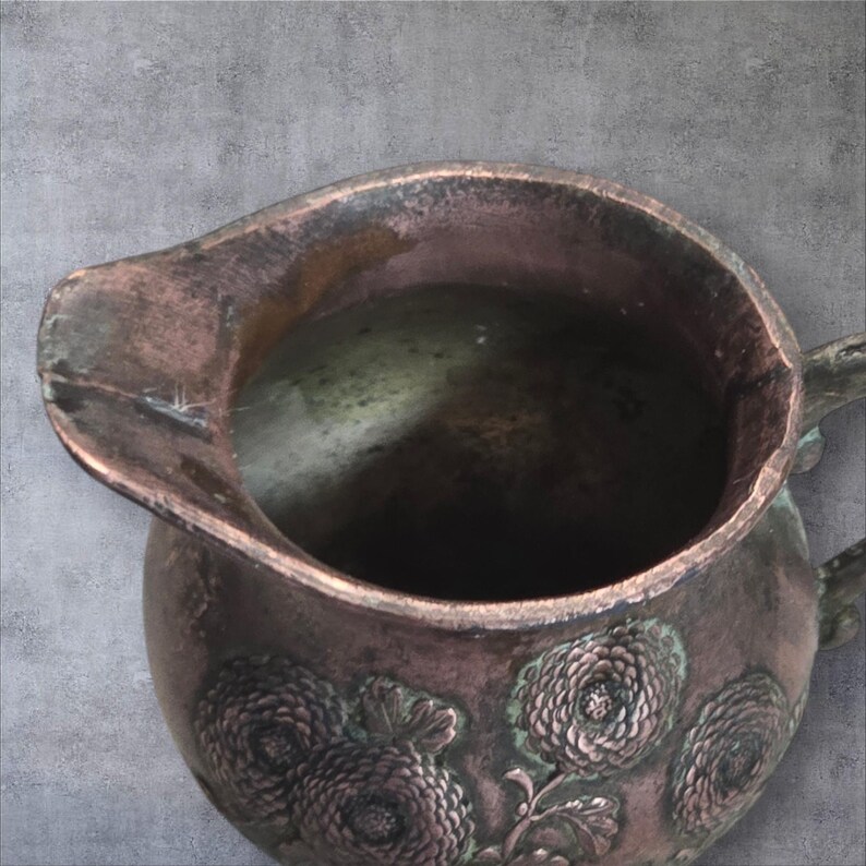 Op de afbeelding: Een vintage koperen kan met een bloemenmotief. De kan heeft een versleten, verweerde uitstraling en is versierd met gegraveerde bloemen.