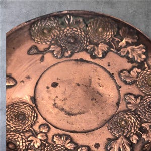 Op de afbeelding: Een koperen dienblad met een reli&euml;fdesign van chrysanten en bladeren. Het dienblad heeft een licht verweerde, verouderde uitstraling.