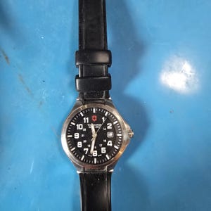 May include: A Victorinox Swiss Army wristwatch with a round silver-tone stainless steel case, a black dial featuring white Arabic numerals for 12 and 24-hour time, a date window at the three o'clock position, and a black rubber watch strap.