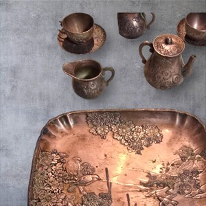 Op de afbeelding: Een koperen theeset met een theepot, roomkan, twee kopjes en schoteltjes en een decoratief dienblad. De set heeft een reli&euml;fmotief van bloemen en een sc&egrave;ne met vogels en bloemen.