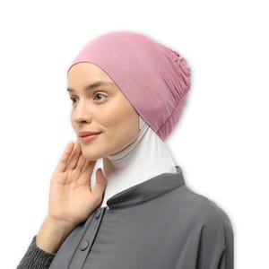 May include: A pink stretchy fabric head covering with a gathered top. The head covering is worn by a person with a white turtleneck shirt and a gray shirt.