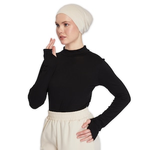May include: A black mock neck shirt with long sleeves and thumbholes. The shirt is made of a soft, stretchy fabric. The model is wearing a beige beanie.