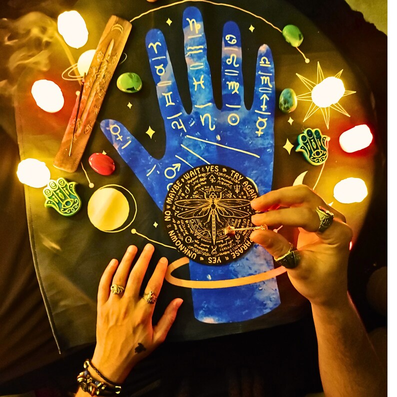 May include: A blue and gold fortune telling board with a hand design and a dragonfly charm. The board has a circular design with the words "Maybe Wait & Yes Try Again" and "Phrase Yes Unknown" written around the edge. The board is surrounded by small stones and a stick of incense.