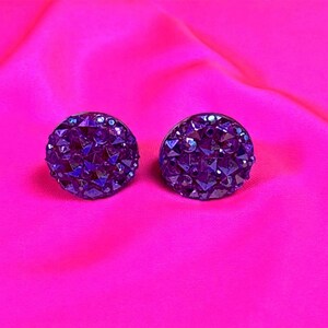 May include: A pair of dark purple stud earrings with a sparkling, faceted surface. The earrings are set against a vibrant pink background, creating a striking contrast. The jewellery is likely made of crystals or rhinestones.