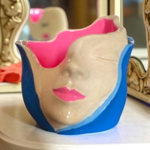 May include: A ceramic vase with a face design. The vase has a blue exterior, a light beige face, and a bright pink interior. The lips are painted pink. The vase has a unique, artistic design.