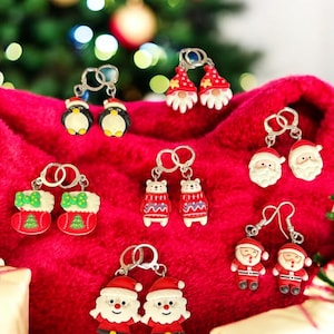 May include: A collection of Christmas-themed earrings, including penguins, gnomes, Santa Claus, polar bears, and stockings. The earrings are red, white, and black, with silver-colored earring hooks. The background is a red fabric with a blurred Christmas tree.