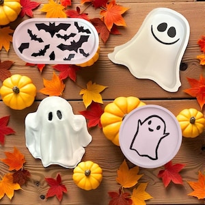 Op de afbeelding: Een verzameling Halloween-thema decoratieve trays en pompoenen op een houten oppervlak. De trays hebben vleermuis- en spookontwerpen. De pompoenen zijn klein en geel, omringd door kleurrijke herfstbladeren.