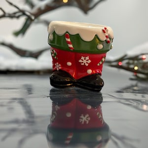 May include: A Christmas-themed ornament in the shape of a Santa's boot. The boot is red with white snowflakes and a green cuff. The top is white with candy cane accents and black shoe. A seasonal holiday item.