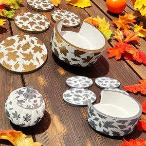 May include: A selection of autumn-themed homeware, including coasters, bowls, and a pumpkin-shaped container. The items feature leaf patterns in gold and grey on a white background. The arrangement is set on a wooden surface with autumn leaves.