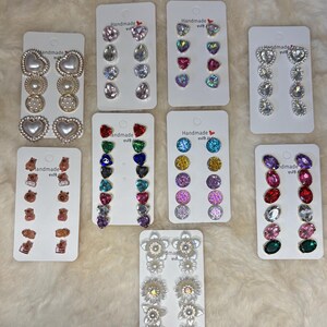 Könnte beinhalten: Eine Sammlung handgefertigter Ohrringe, präsentiert auf weißen Karten. Stile umfassen Perlenherzen, klare und farbige Edelsteine, herzförmige Ohrstecker und Blumenmuster. Die Karten tragen den Text "Handmade with love."