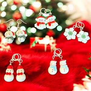 Könnte beinhalten: Eine Sammlung von Ohrringen zum Thema Weihnachten. Die Ohrringe zeigen Schneemann- und Schneeflocken-Designs in Rot, Weiß und Blau. Die Ohrringe werden auf einer roten, flauschigen Oberfläche mit einem verschwommenen Weihnachtsbaum-Hintergrund präsentiert.