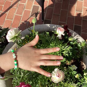 Könnte beinhalten: Ein grünes und weißes Perlenarmband mit kleinen goldenen Perlen und grünen Kleeblatt-Anhängern. Das Armband befindet sich am Handgelenk einer Person, deren Hand über einem Topf mit weißen und rosa Blüten ausgestreckt ist.