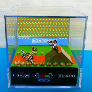 Puede incluir: Un diorama 3D del clásico videojuego de NES 'Excitebike'. El diorama presenta un motociclista en miniatura saltando sobre una rampa, con un fondo pixelado y una pantalla de puntuación que muestra '3RD 1:24:00 TEMP 0:06:81 TIME'.