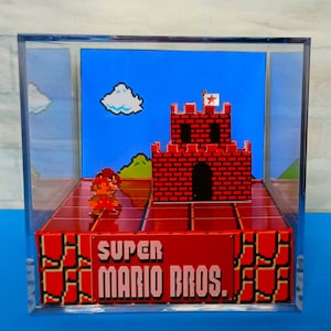Puede incluir: Una vitrina de acrílico transparente con un nivel en miniatura de Super Mario Bros. de 8 bits en su interior. El nivel presenta un castillo de ladrillos rojos con una bandera en la cima, una colina verde y una pequeña figura de Mario de pie sobre una plataforma de ladrillos rojos. La base de la vitrina tiene el texto "SUPER MARIO BROS." impreso en ella.