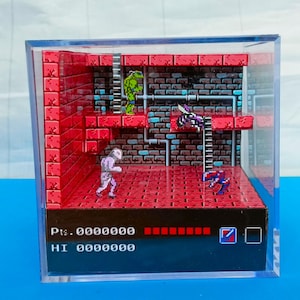 Puede incluir: Un diorama de una escena de videojuego clásico, con personajes pixelados y un entorno de ladrillos rojos y marrones. La escena está encerrada en un cubo de acrílico transparente. La puntuación y la barra de salud del juego son visibles en la parte inferior.