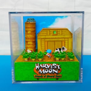 Könnte beinhalten: Ein durchsichtiger, würfelförmiger Präsentationskoffer aus Kunststoff mit einem Miniatur-Diorama einer Farm-Szene aus dem Videospiel Harvest Moon. Das Diorama zeigt einen Silo, einen Stall, eine Kuh, einen Bauern und ein Feld mit Feldfrüchten. Der Text "Harvest Moon Friends of Mineral Town" ist auf der Basis des Präsentationskoffers aufgedruckt.