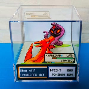Może przedstawiać: Trójwymiarowa dioramka przedstawiająca walkę Pokémonów między Charizardem a Arbokiem. Charizard to czerwony i pomarańczowy smokopodobny Pokémon z dużymi skrzydłami. Arbok to fioletowo-czarny wężopodobny Pokémon. Dioramka jest umieszczona na niebieskim tle i zawiera zieloną trawiastą przestrzeń. Tekst na dioramce brzmi "Co zrobi CHARIZARD?" i "WALCZ POKÉMON UCIEKAJ".