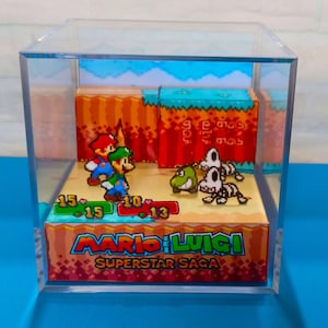 May include: A diorama of a scene from the video game "Mario & Luigi: Superstar Saga" inside a clear acrylic box. The scene features Mario and Luigi facing enemies, with health bars displayed. The game's logo is at the bottom.