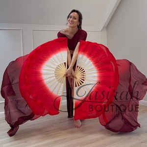 May include: A pair of dance fans featuring a bamboo frame and a fabric canopy that transitions from a white center to a vibrant red outer edge, with long, flowing dark burgundy silk extensions. The text Jasirah BOUTIQUE is visible.