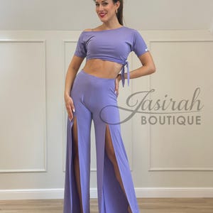 May include: A lavender two-piece outfit featuring a crop top with an off-the-shoulder design and tie detail, paired with wide-leg pants with high slits. The outfit is made of a soft, flowing fabric. The text "Jasirah Boutique" is visible.