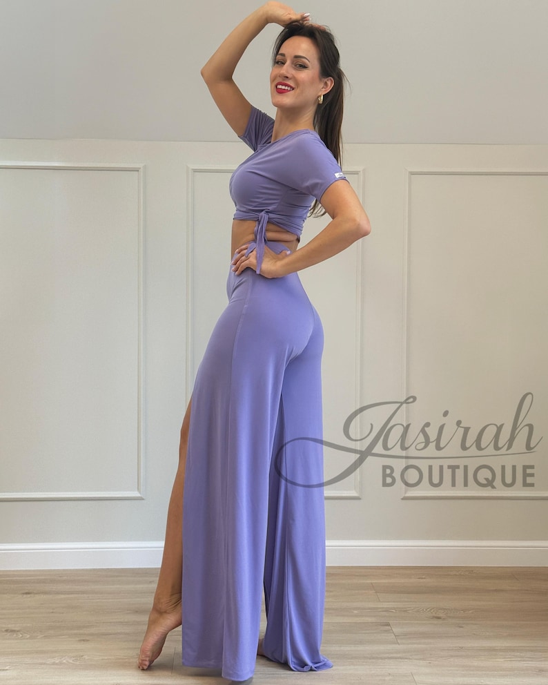 May include: A lavender two-piece outfit, featuring a short-sleeved crop top with tie details and high-waisted, wide-leg pants with a side slit. The outfit is made of a soft, flowing fabric. The boutique's logo is visible in the bottom right corner.