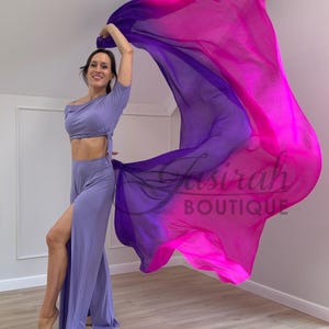 Könnte beinhalten: Ein lavendelfarbenes zweiteiliges Outfit bestehend aus einem kurzärmeligen bauchfreien Oberteil und einer hoch taillierten Hose mit Seitenschlitz, kombiniert mit einem großen, fließenden Seidenschleier mit einem Farbverlauf von Dunkelviolett zu Pink.