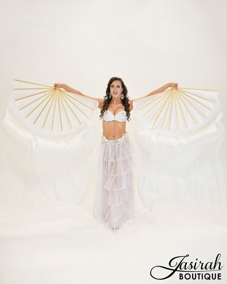 Puede incluir: Una mujer con un traje de danza del vientre blanco con un sujetador enjoyado y una falda en capas, sosteniendo grandes abanicos blancos y dorados. Los abanicos est&aacute;n muy extendidos, creando un efecto dram&aacute;tico. El logotipo de Jasirah Boutique est&aacute; en la esquina inferior derecha.