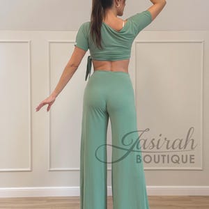 May include: A sage green two-piece outfit featuring a crop top with short sleeves and a tie closure, paired with wide-leg pants. The outfit is made of a soft, flowing fabric. The logo "Jasirah Boutique" is visible.