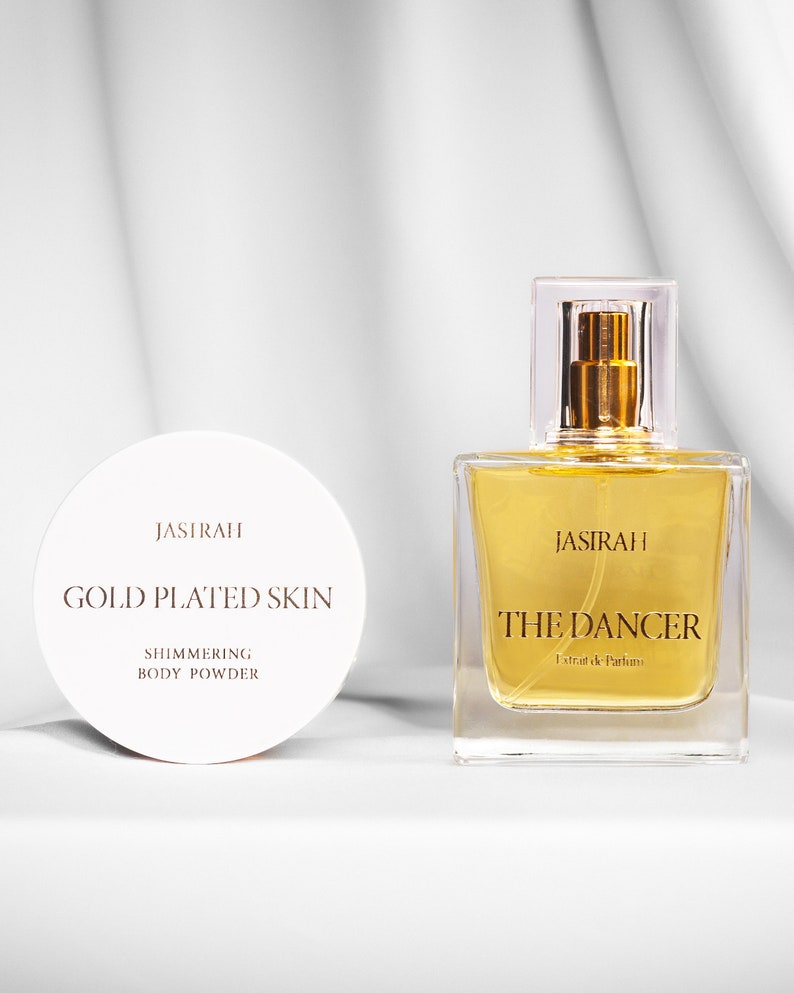 May include: A cosmetic product set featuring a round, white container labeled "JASIRAH GOLD PLATED SKIN SHIMMERING BODY POWDER" and a square perfume bottle with "JASIRAH THE DANCER Extrait de Parfum" text.