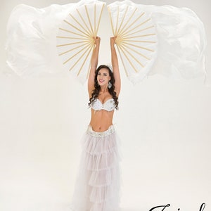 Puede incluir: Una mujer con un traje blanco de danza del vientre con un sujetador enjoyado y una falda escalonada, sosteniendo dos abanicos blancos y dorados. Los abanicos est&aacute;n abiertos, con tela fluida. La imagen est&aacute; sobre un fondo blanco. El logo "Jasirah Boutique" est&aacute; en la esquina inferior derecha.