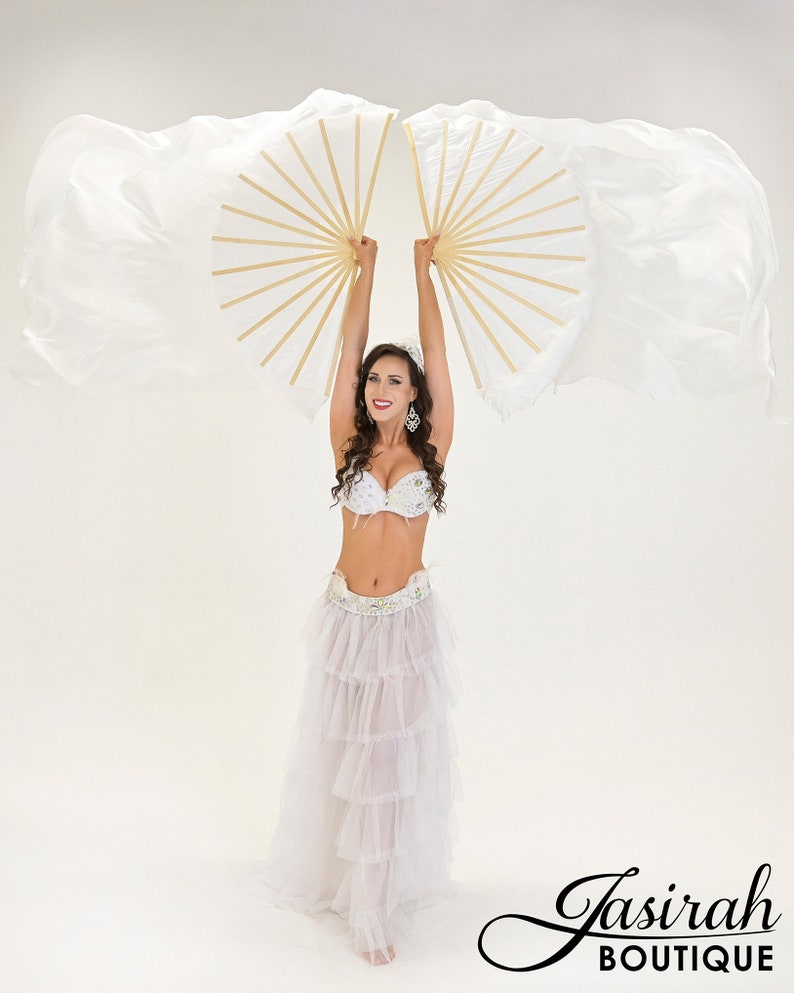 Puede incluir: Una mujer con un traje blanco de danza del vientre con un sujetador enjoyado y una falda escalonada, sosteniendo dos abanicos blancos y dorados. Los abanicos est&aacute;n abiertos, con tela fluida. La imagen est&aacute; sobre un fondo blanco. El logo "Jasirah Boutique" est&aacute; en la esquina inferior derecha.