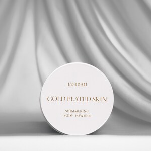 May include: A white, round container of shimmering body powder. The container has gold lettering that reads "JASIRAH GOLD PLATED SKIN SHIMMERING BODY POWDER". The background is a white, draped fabric.