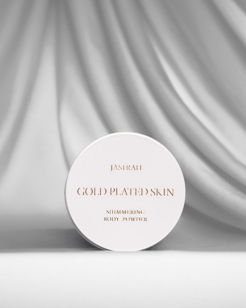 May include: A white, round container of shimmering body powder. The container has gold lettering that reads "JASIRAH GOLD PLATED SKIN SHIMMERING BODY POWDER". The background is a white, draped fabric.