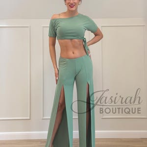 May include: Sage green two-piece outfit. The top is an off-the-shoulder crop top with a tie detail. The matching pants have high slits on the sides. The outfit is made of a soft, flowing fabric. The text "Tasirah Boutique" is visible.