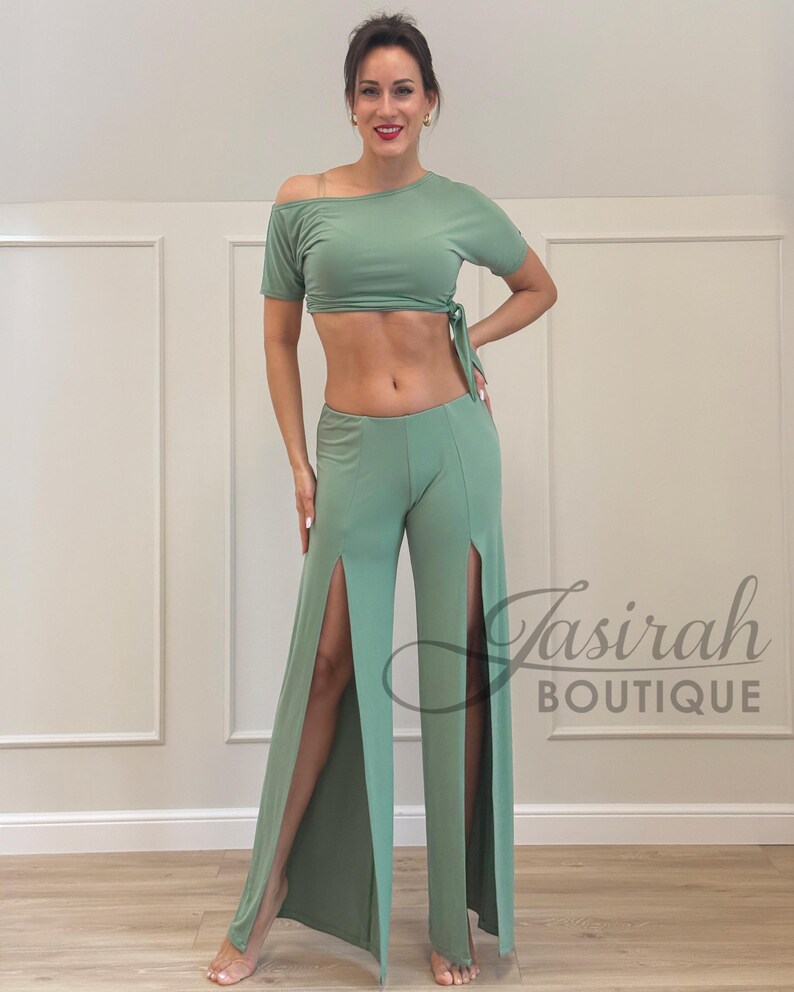 May include: Sage green two-piece outfit. The top is an off-the-shoulder crop top with a tie detail. The matching pants have high slits on the sides. The outfit is made of a soft, flowing fabric. The text "Tasirah Boutique" is visible.