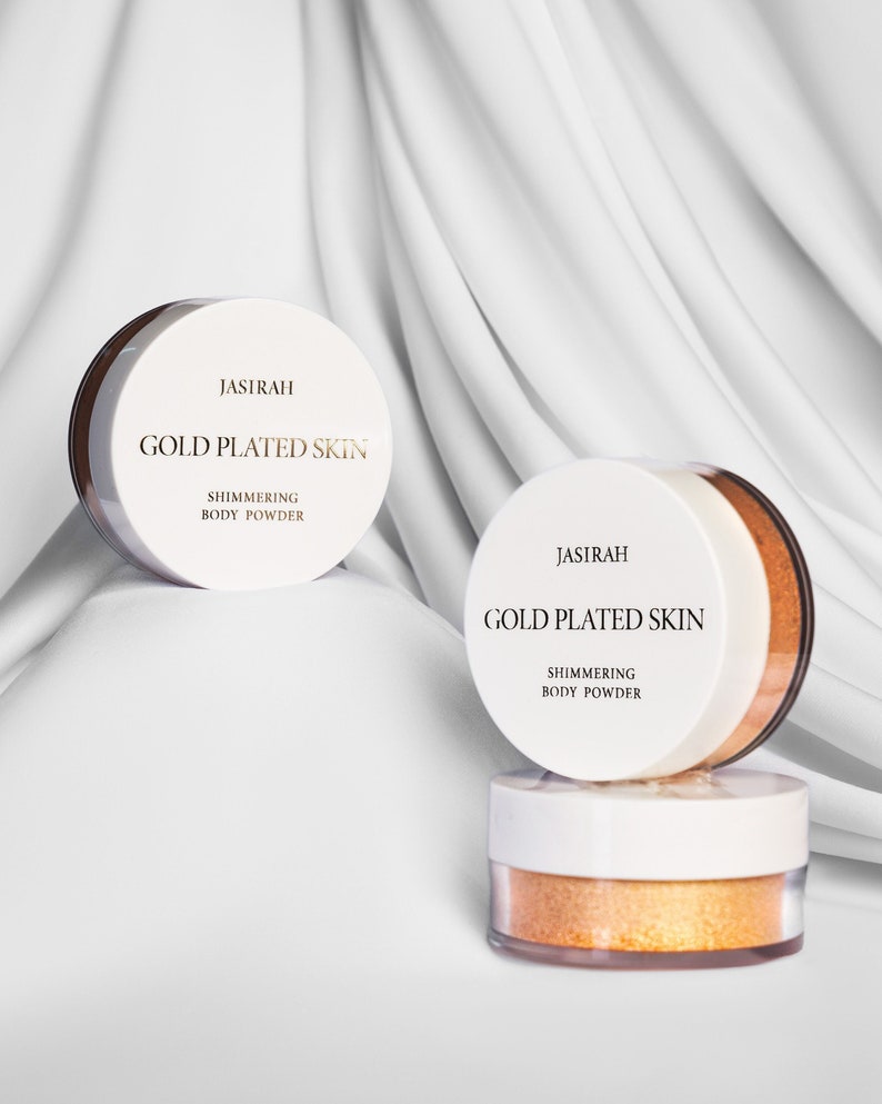 May include: Three jars of shimmering body powder with white lids and gold lettering that reads "JASIRAH GOLD PLATED SKIN SHIMMERING BODY POWDER." The powder inside is a bright gold color. The jars are arranged on a white fabric background.