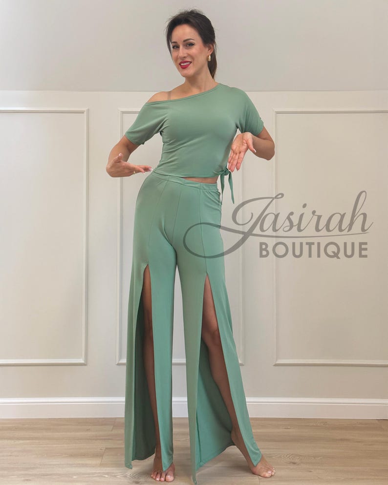 May include: Sage green two-piece outfit. The top is an off-the-shoulder style with a tie at the waist. The pants are wide-legged with high slits on the sides. The outfit is from Jasirah Boutique.