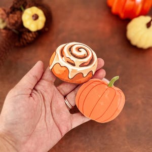 May include: Two flat, circular decorative items featuring illustrations of a cinnamon roll with white icing and a solid orange pumpkin with a green stem. The cinnamon roll has brown swirls and a tan base, while the pumpkin has vertical lines.