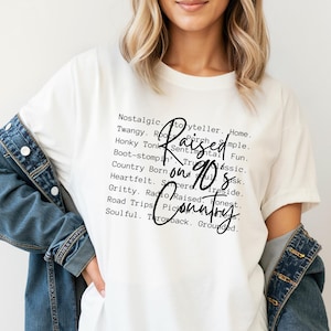 Può includere: T-shirt color crema con la scritta "Raised on 90s Country" in nero. La maglietta presenta un elenco di parole legate alla musica country in un carattere nero più piccolo. Una giacca di jeans è indossata sulle spalle.