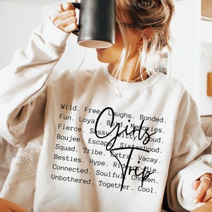 May include: A cream-colored sweatshirt with the words "Girls Trip" in a script font. The sweatshirt also has a list of words such as "Wild", "Free", "Fun", and "Besties". A black mug is held up to the face.